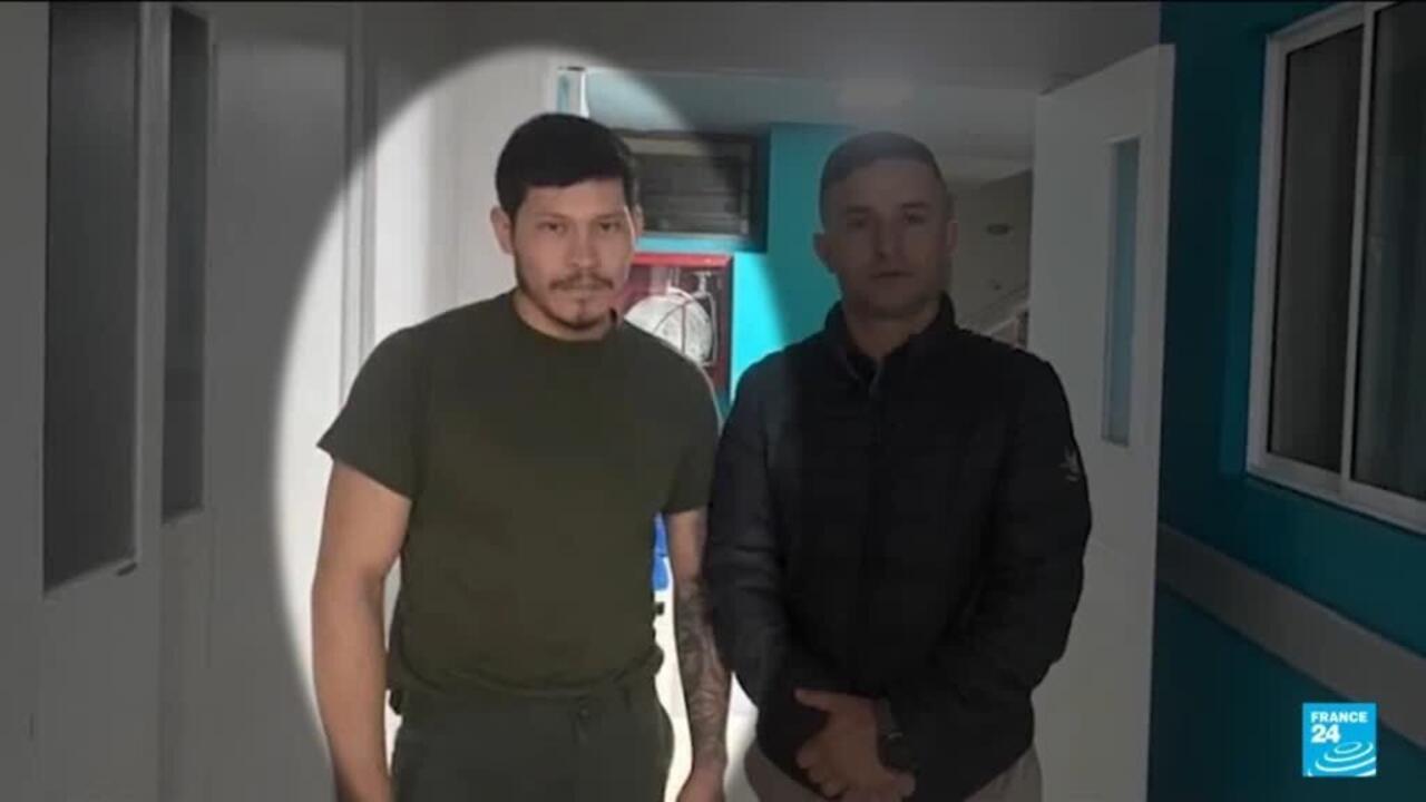 Two men stand indoors, one illuminated by a spotlight, with a France 24 logo visible.