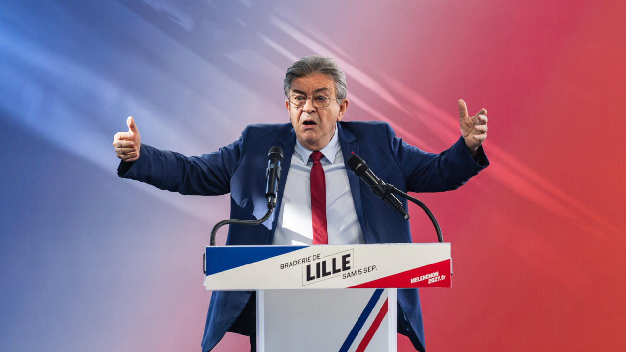A gray-haired man in a navy suit gestures behind a lectern with microphones against a blue and red backdrop.
