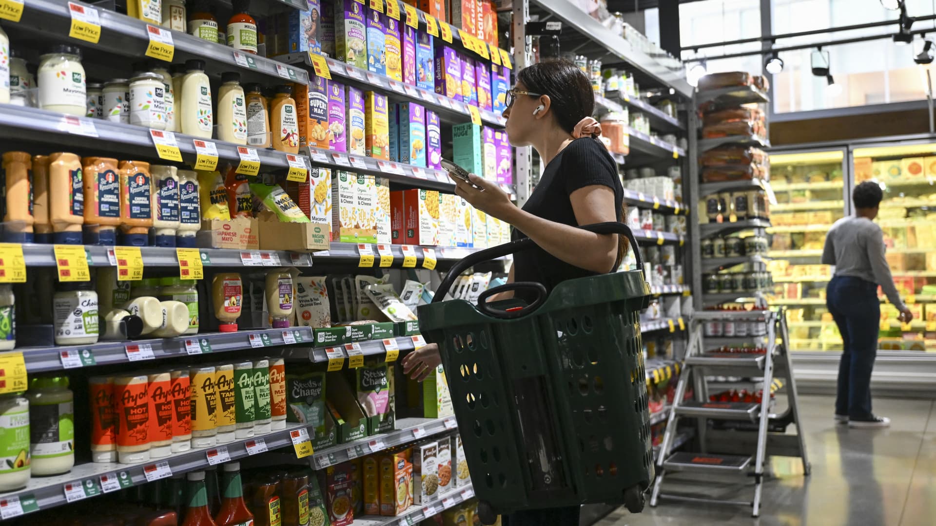 Shopper examines products with a basket in a brightly lit grocery store aisle.