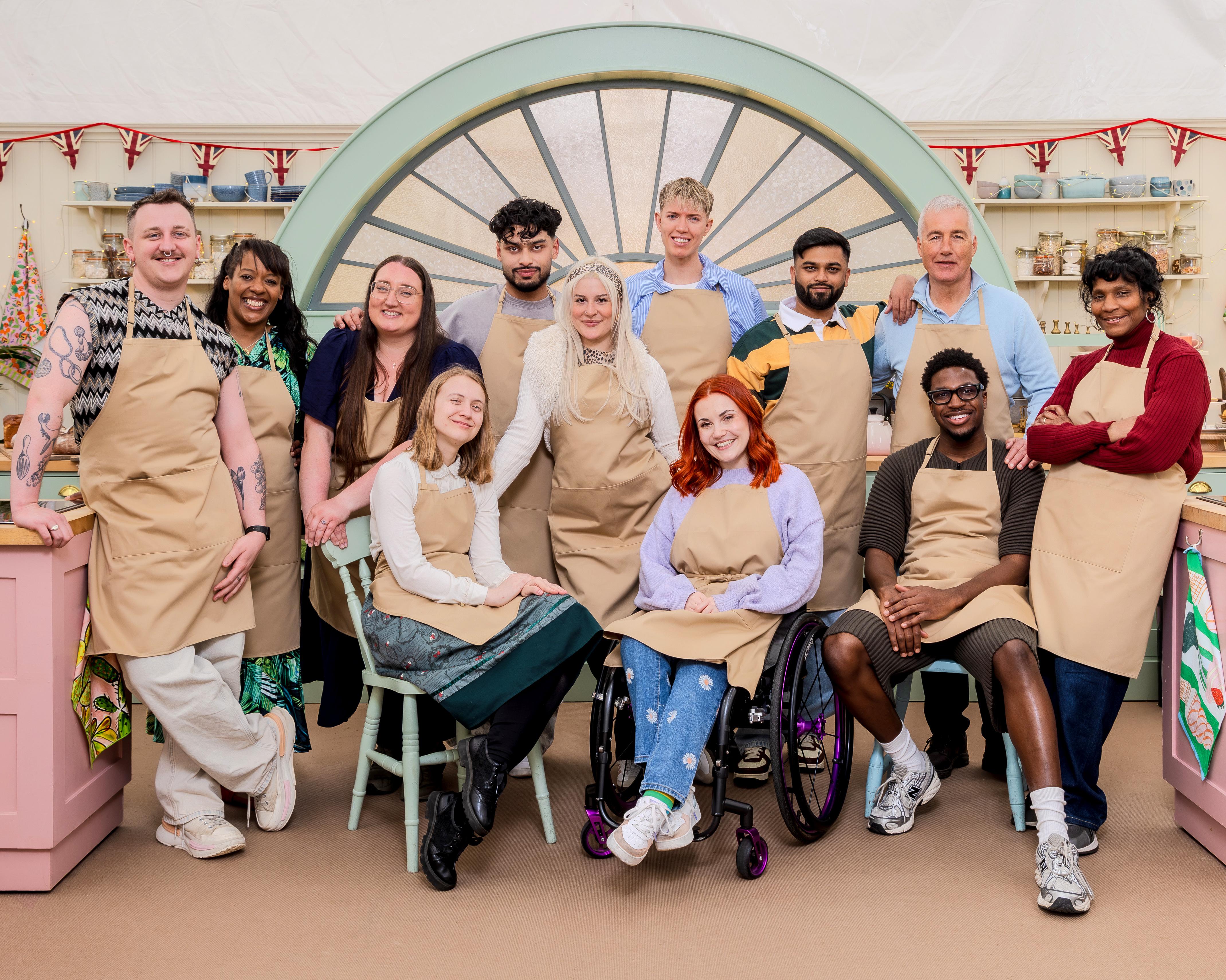 Group of Bake Off contestants and presenters posing together in a brightly decorated bakery setting