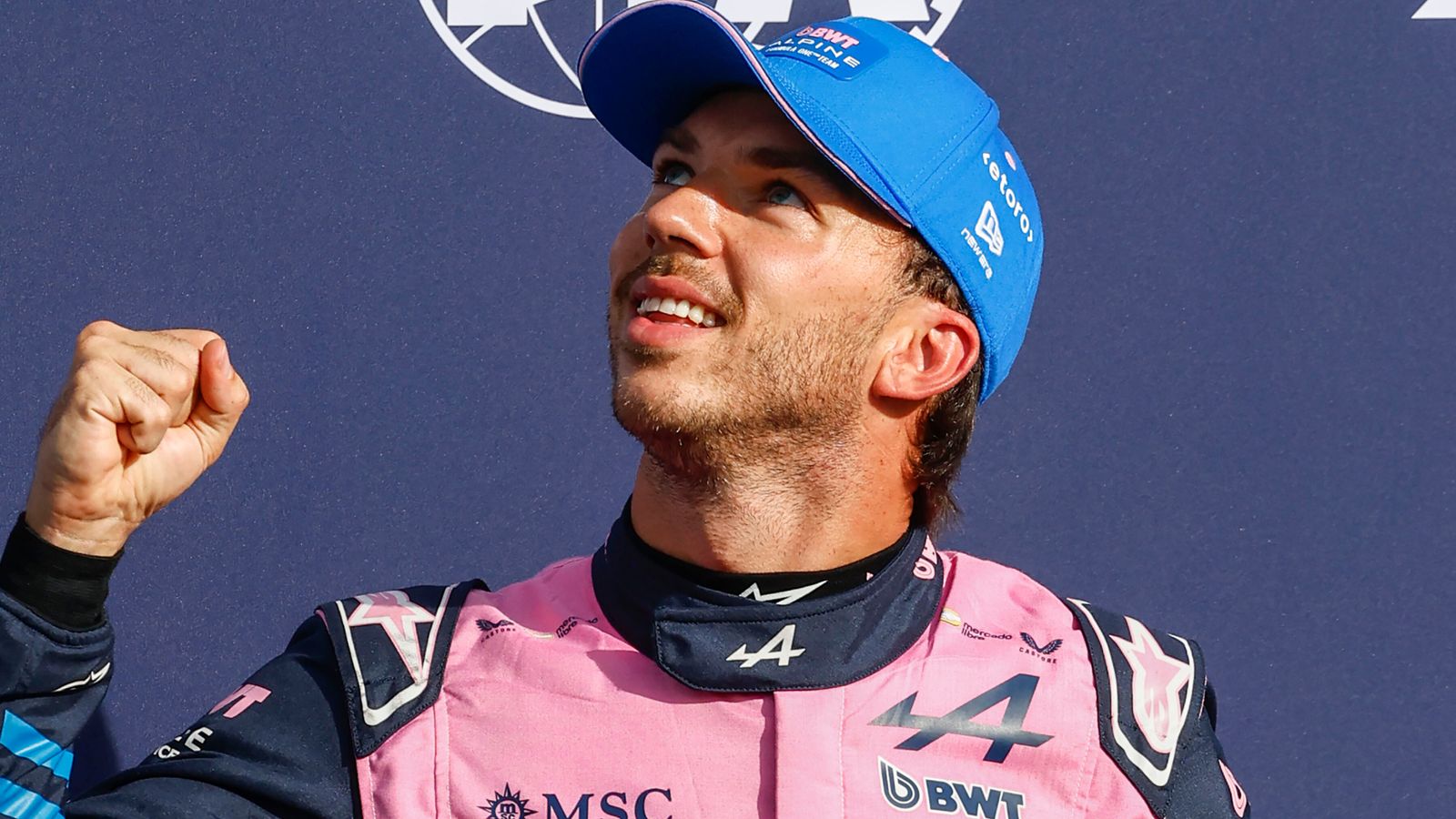 Racing driver in pink and navy suit raises a clenched fist while wearing a blue cap