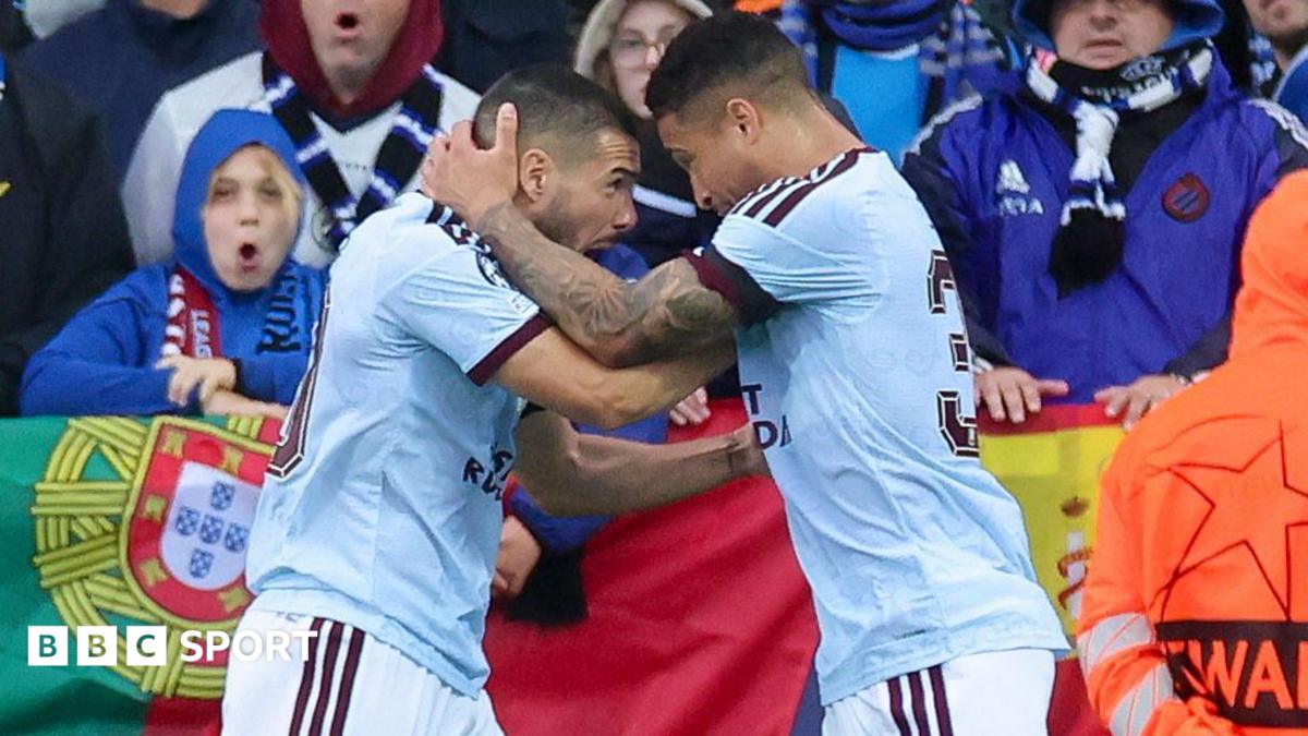 Two footballers in white kits embrace near the touchline as fans watch behind them.