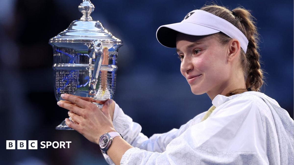 Tennis player holding a large silver trophy while wearing a white visor and jacket