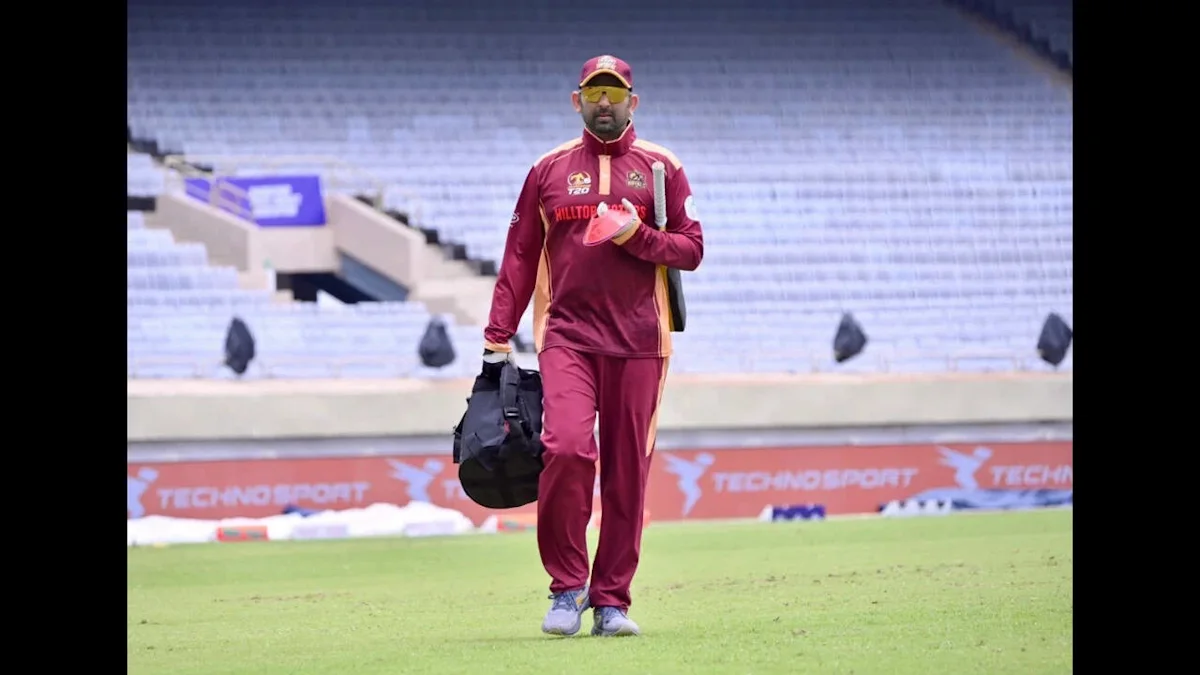 A cricketer in a maroon uniform walks across a grass field carrying sports bags.