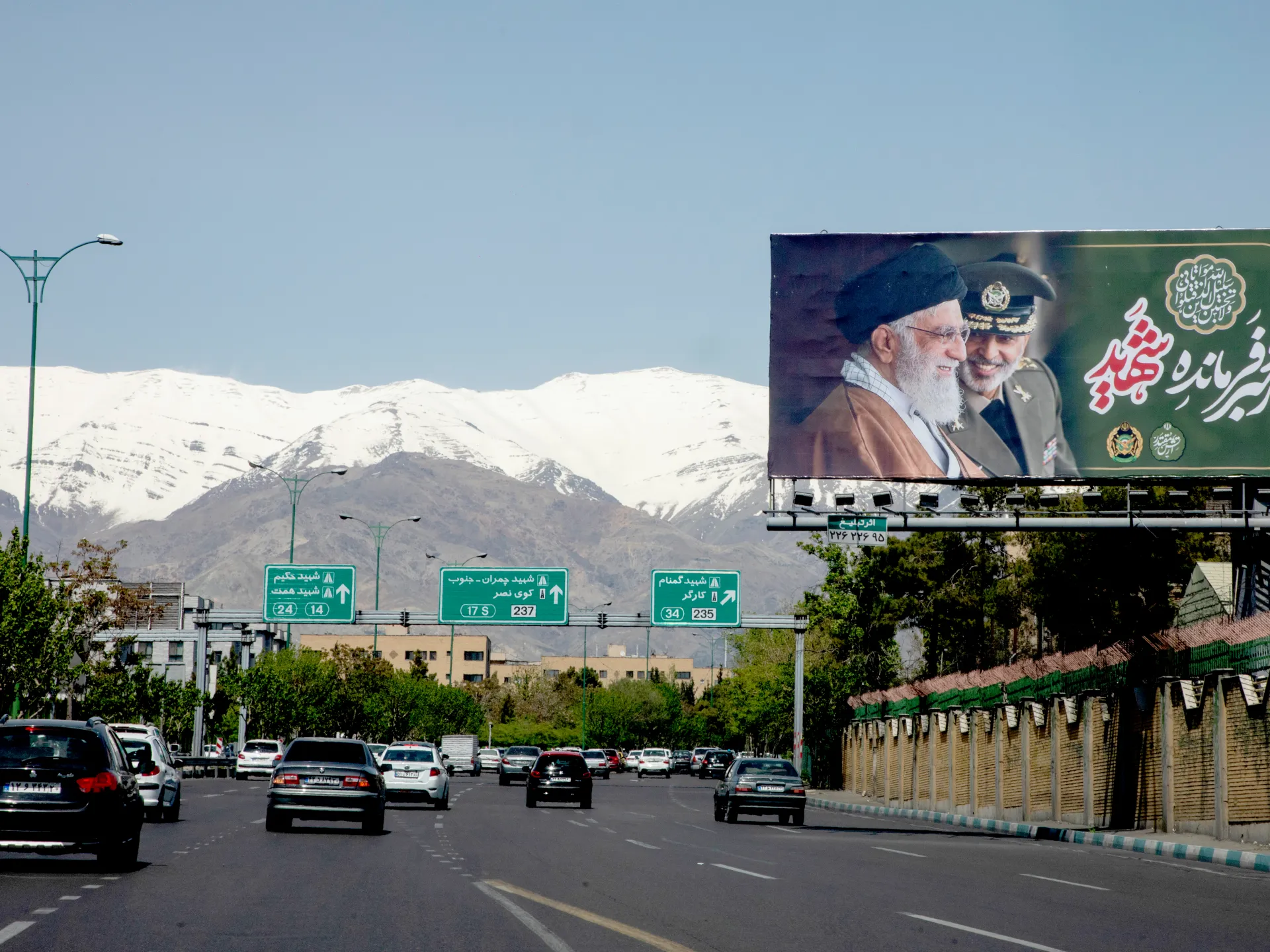 Traffic moves along a broad highway beneath snow-capped mountains and a large Persian-language billboard.