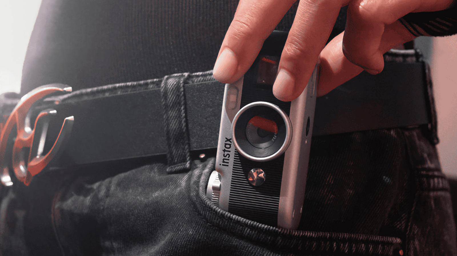 A hand places a compact Instax camera into the front pocket of dark jeans.