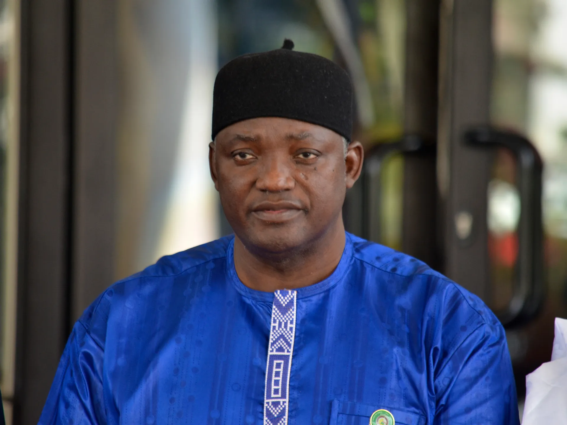 Man wearing a blue traditional outfit and black cap, seated indoors and facing the camera.