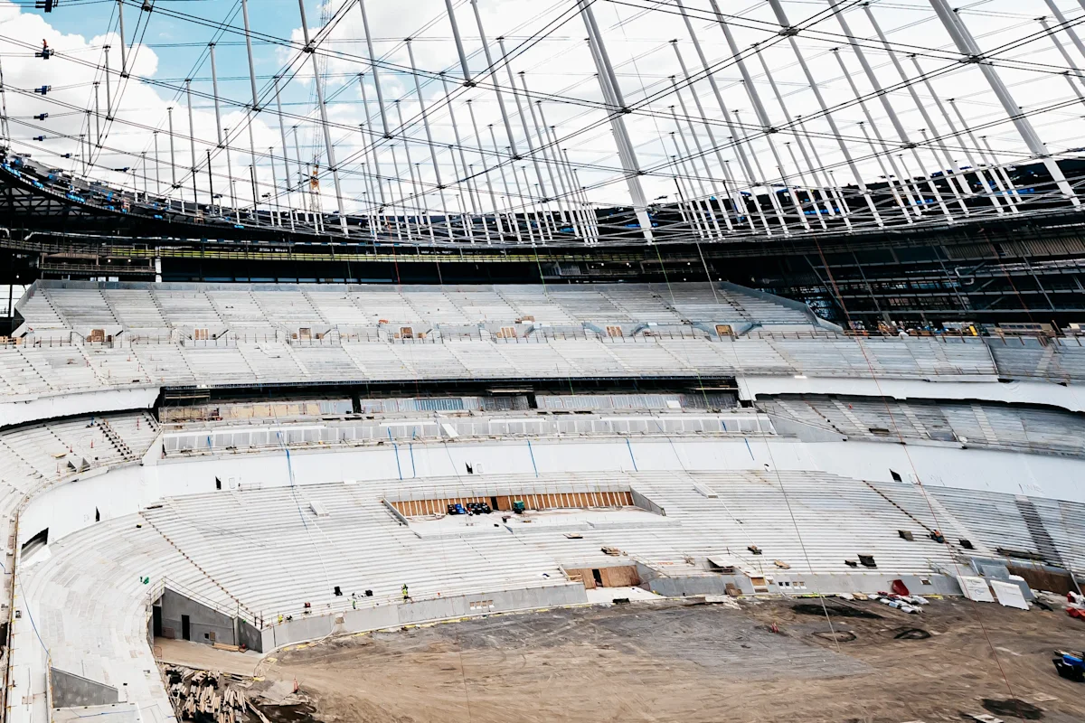 Under-construction stadium interior with empty concrete seating, exposed roof supports, and construction equipment.