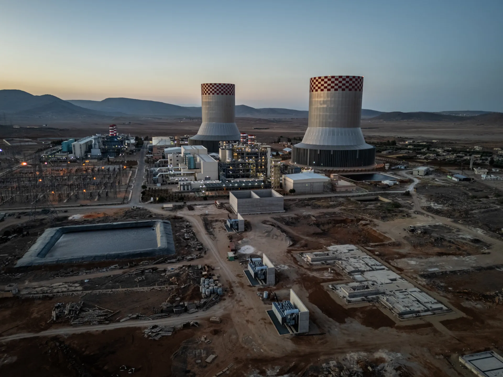 Aerial view of an industrial power facility with two cooling towers, construction grounds, and surrounding hills at dusk.