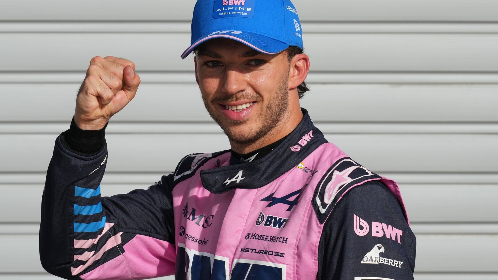 Formula One driver in pink and navy racing suit raises a clenched fist while smiling