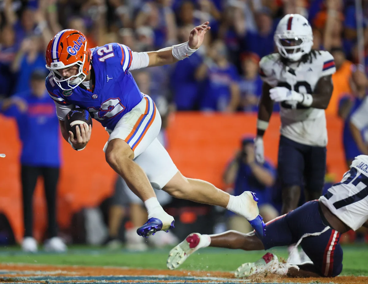 Florida quarterback in blue and orange leaps forward with the football as defenders attempt a tackle