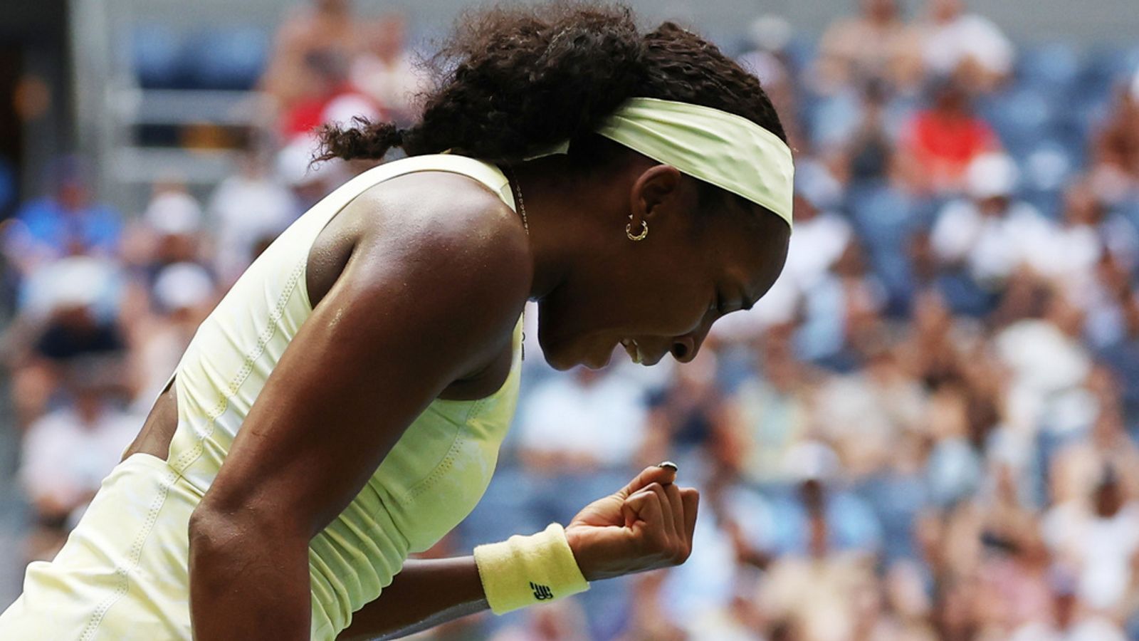 Tennis player in pale outfit bends forward and clenches fist before a blurred crowd.