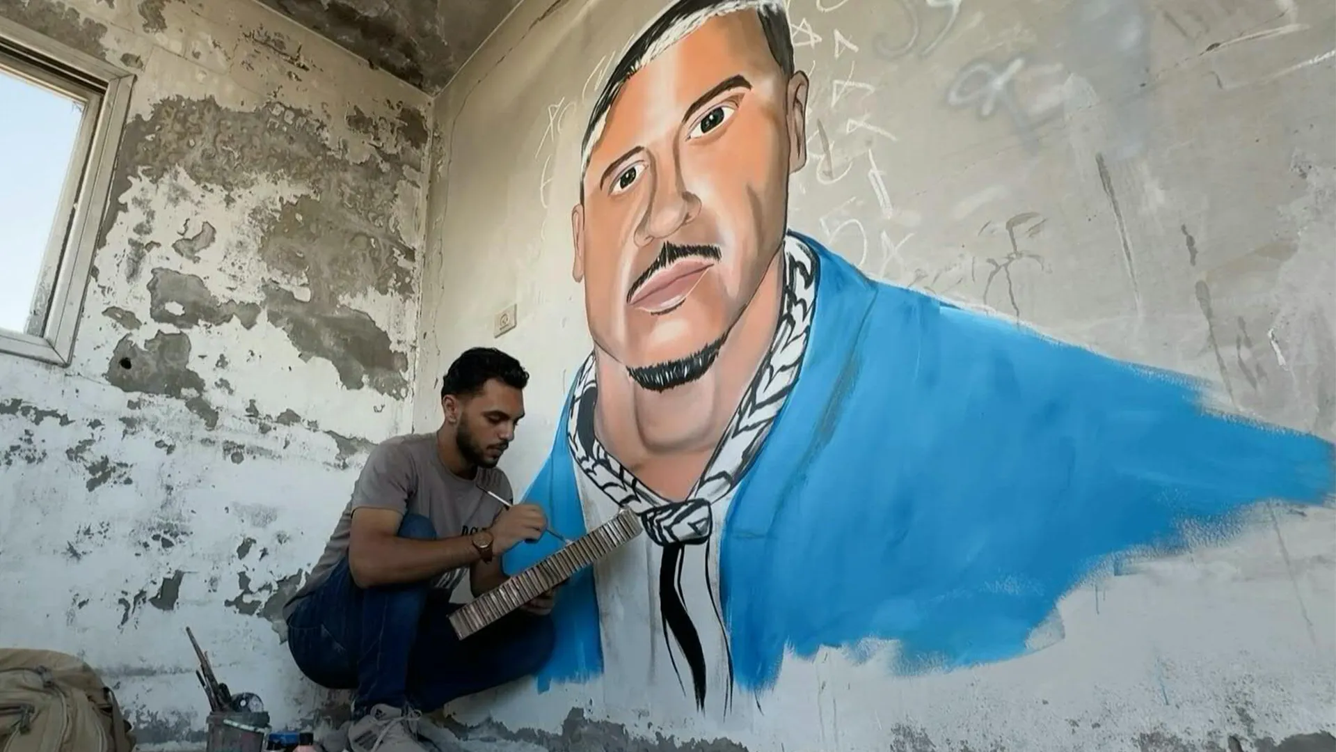 Artist paints a large blue-jacketed portrait mural inside a weathered, partially stripped room.