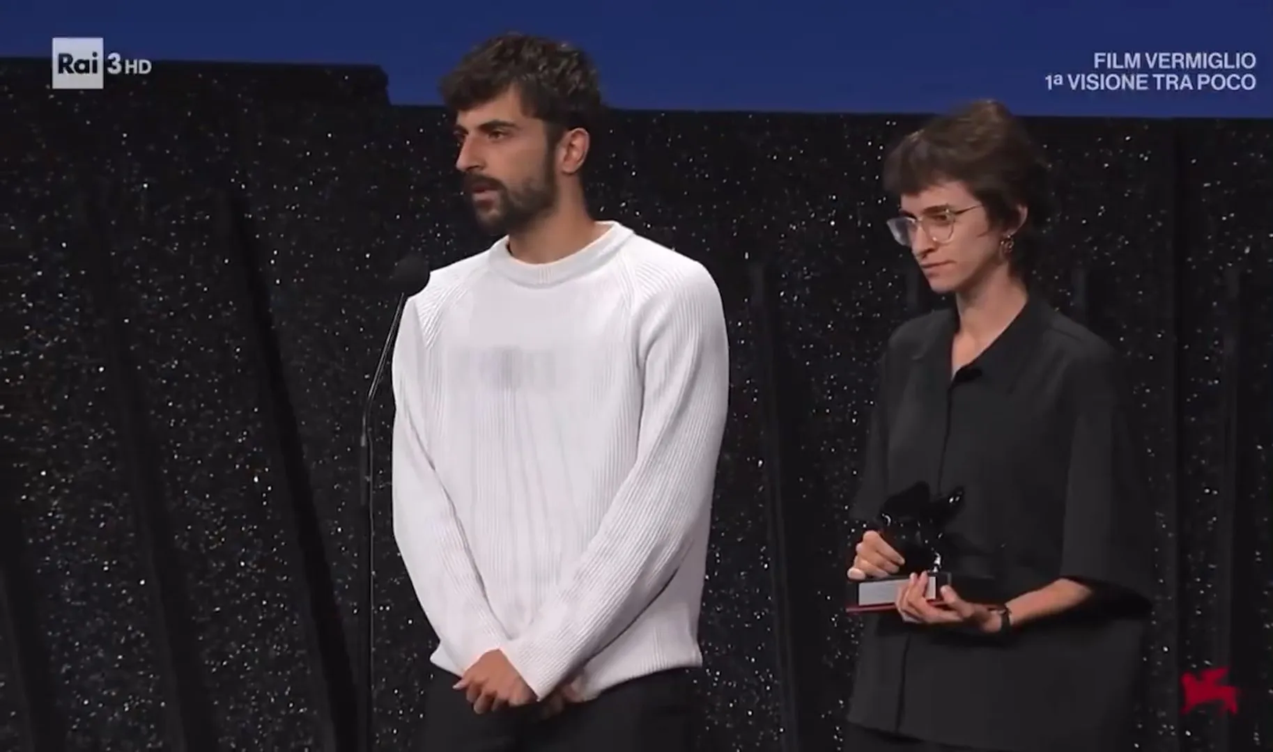 Man in white sweater stands beside woman holding a trophy during an award presentation