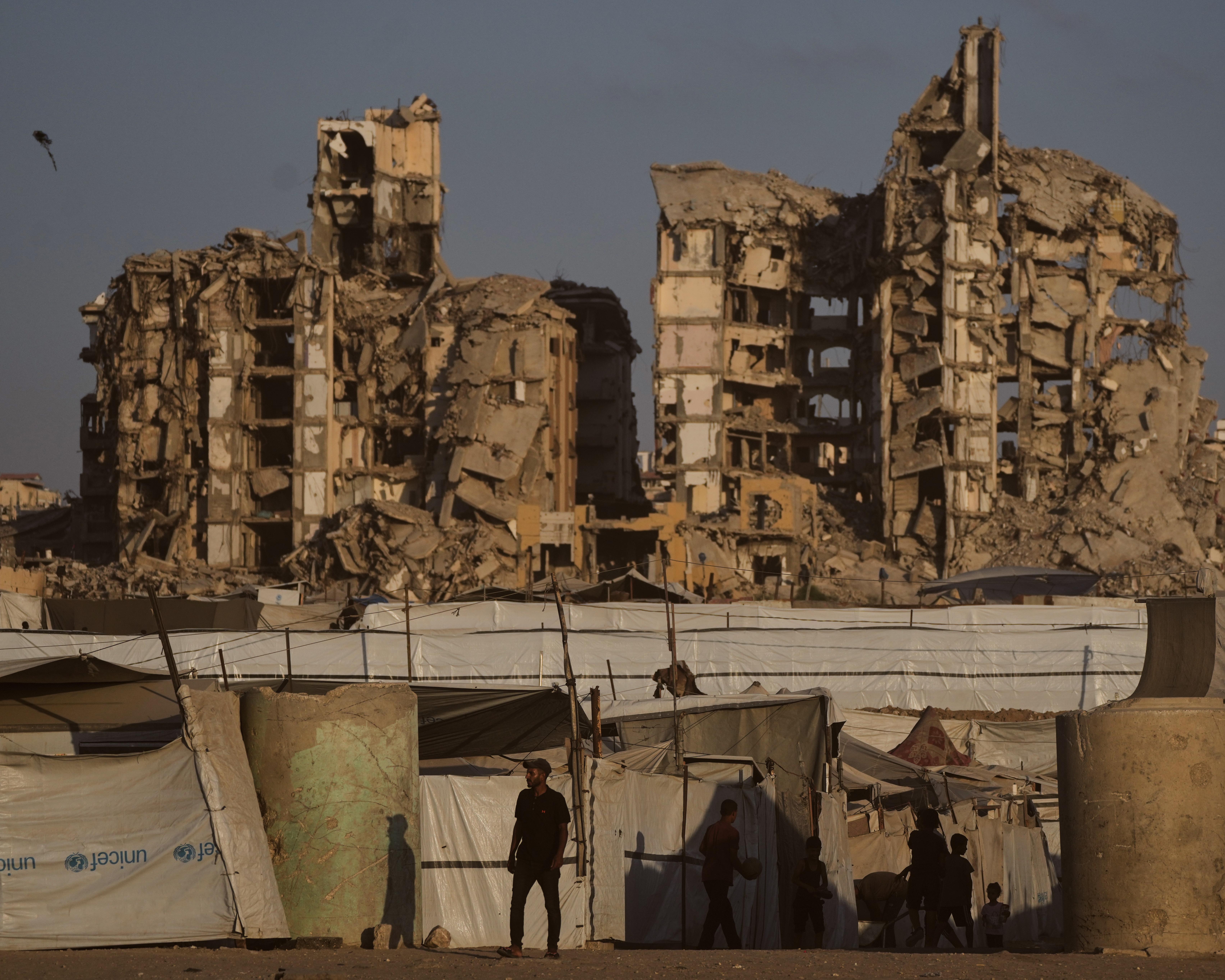 People stand among makeshift tents in front of heavily damaged, partially collapsed buildings.
