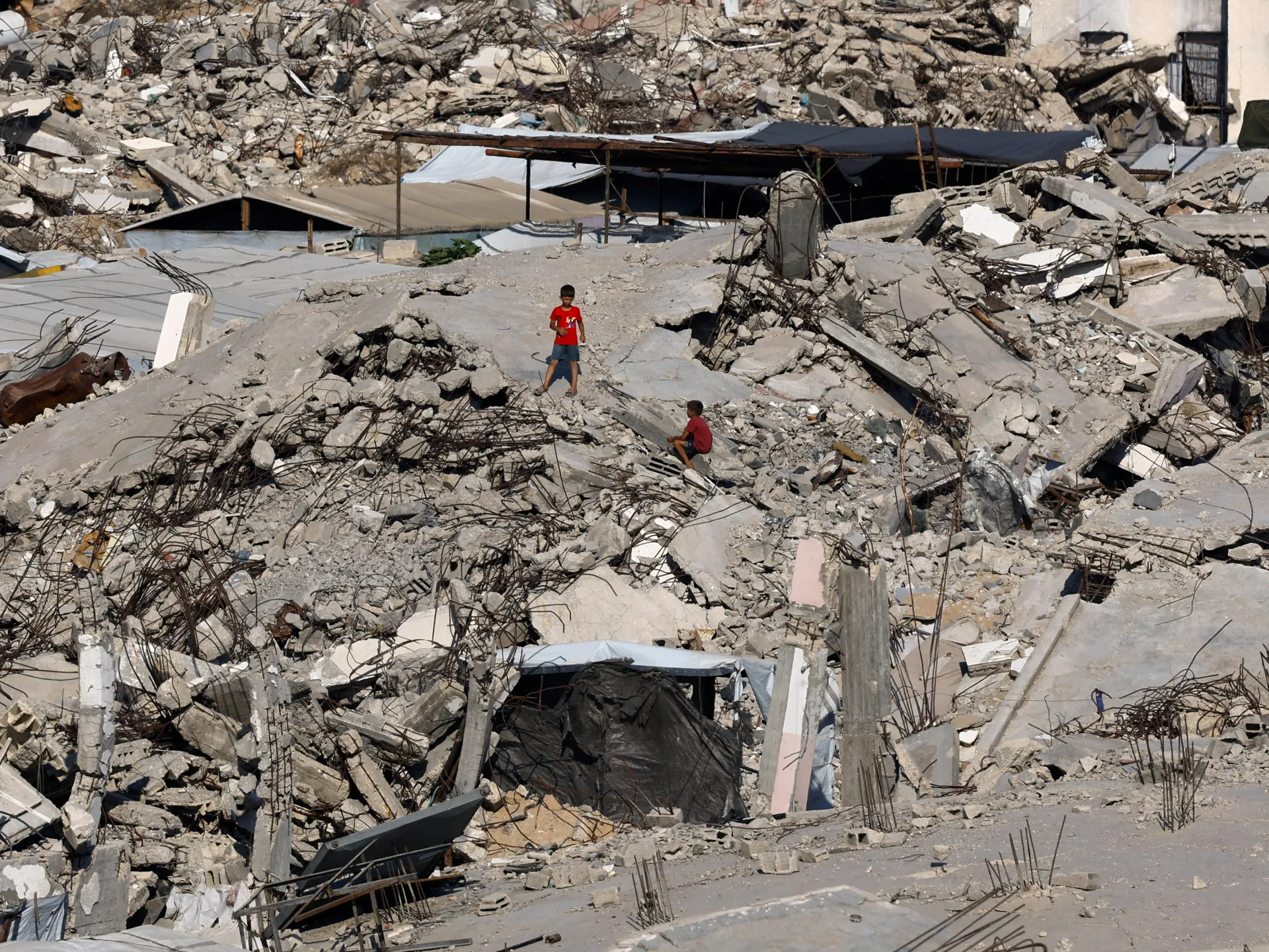 Two children stand amid the rubble of a collapsed residential building and exposed reinforcement bars.