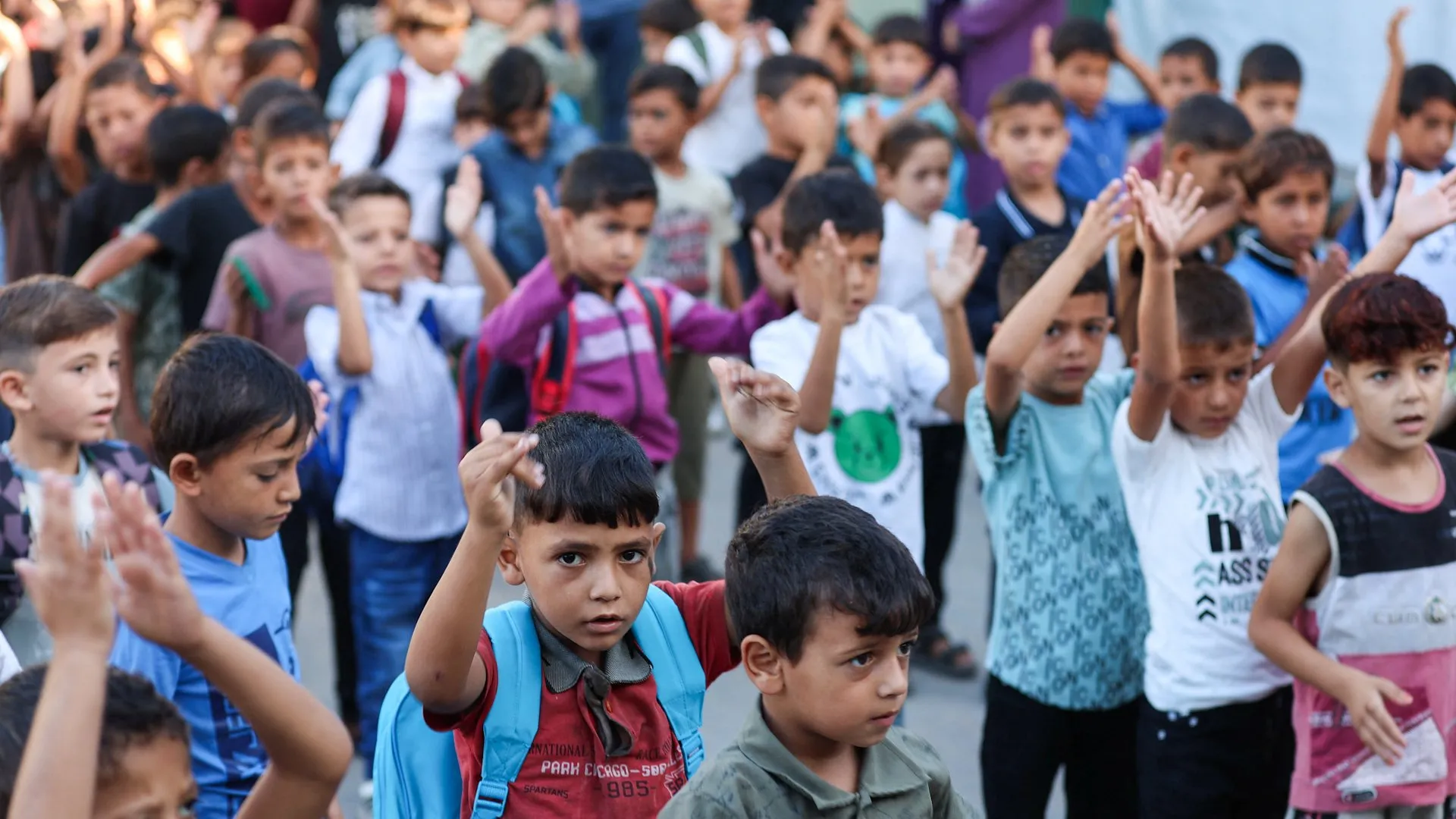 Children with backpacks raise their hands in a crowded outdoor gathering.
