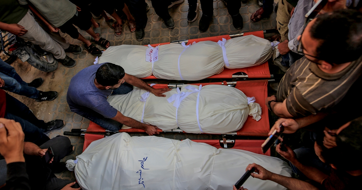 People gather around three shrouded bodies placed on red stretchers.