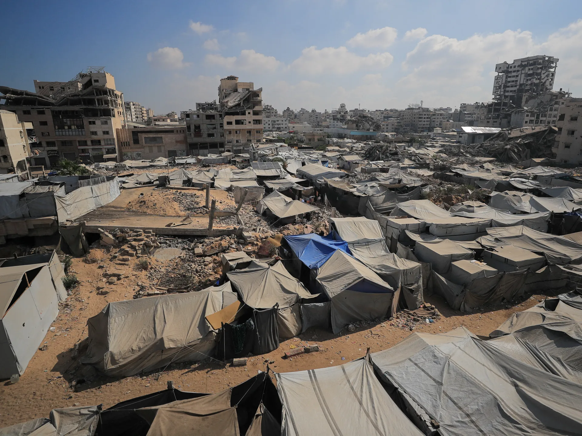 A dense field of white and gray tents fills a sandy area beneath damaged buildings and a hazy sky.