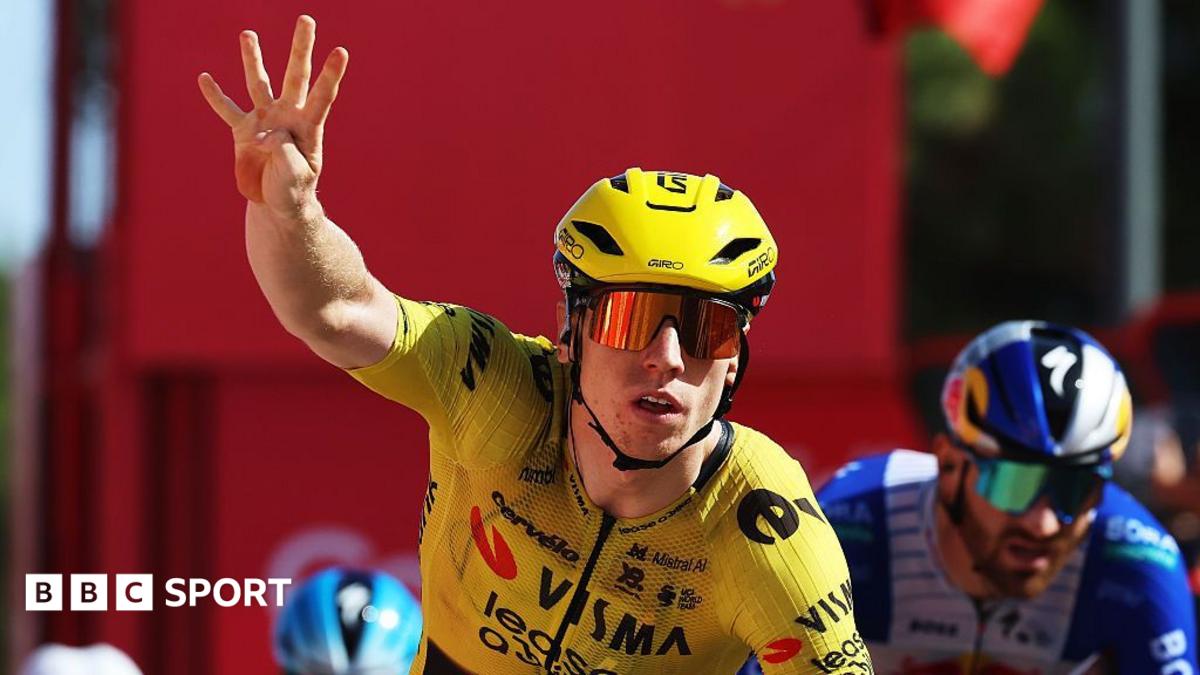 Cyclist in yellow kit raises four fingers while riding ahead of another cyclist.