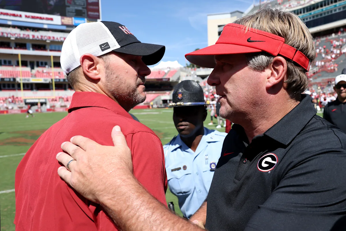Two football coaches converse on the field during a sunny game day, with a security officer behind them.