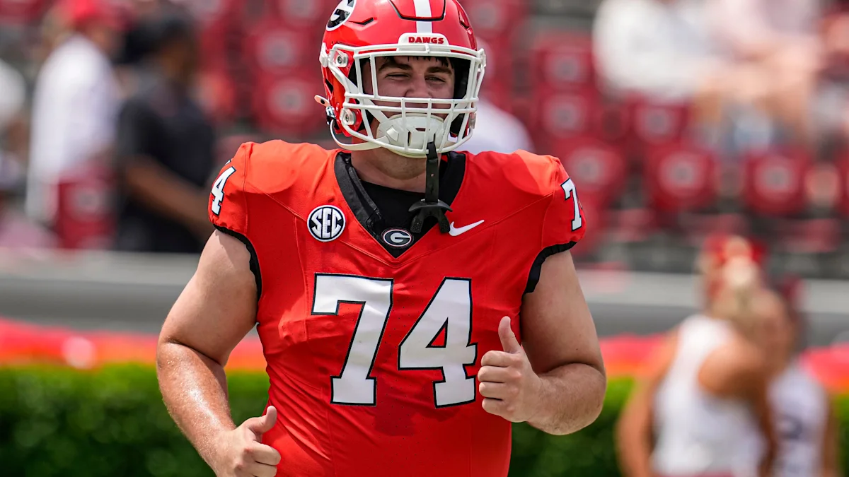Football player in a red number 74 jersey and helmet gives two thumbs up on a field.