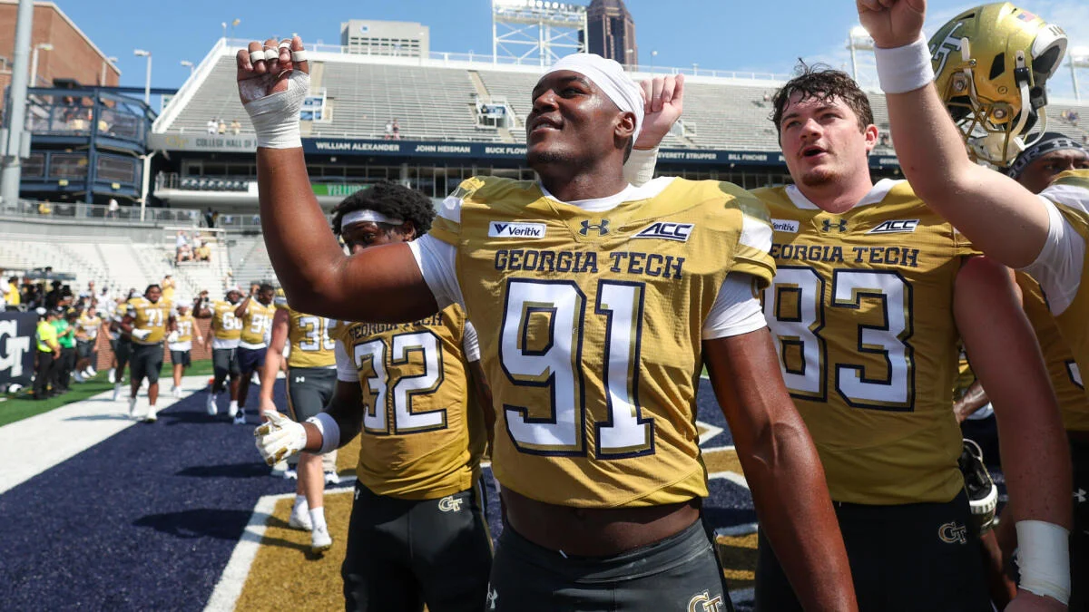 Georgia Tech football players in gold jerseys walk together on the field, raising their arms.