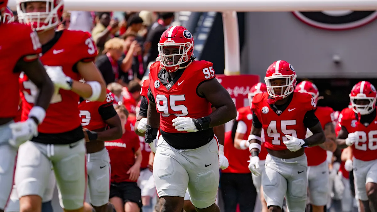Georgia football players in red jerseys and white pants run onto the field before a game.