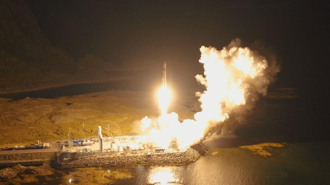 Rocket launches at night from a coastal facility, illuminating smoke, water and surrounding terrain.