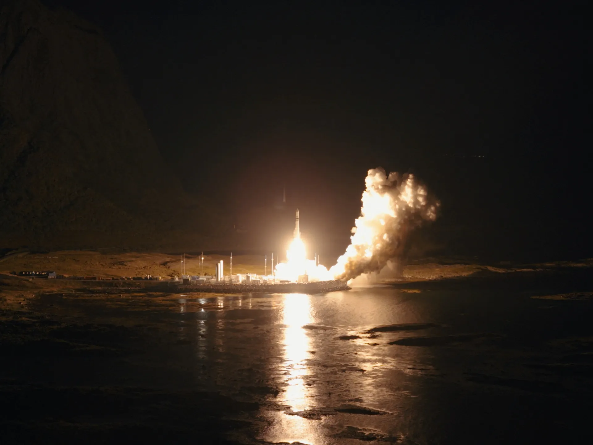 Rocket launches at night from a coastal facility, illuminating smoke, rugged terrain and calm water.