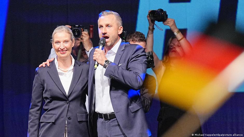 Two politicians stand onstage as one speaks into a microphone before photographers and colorful flags.