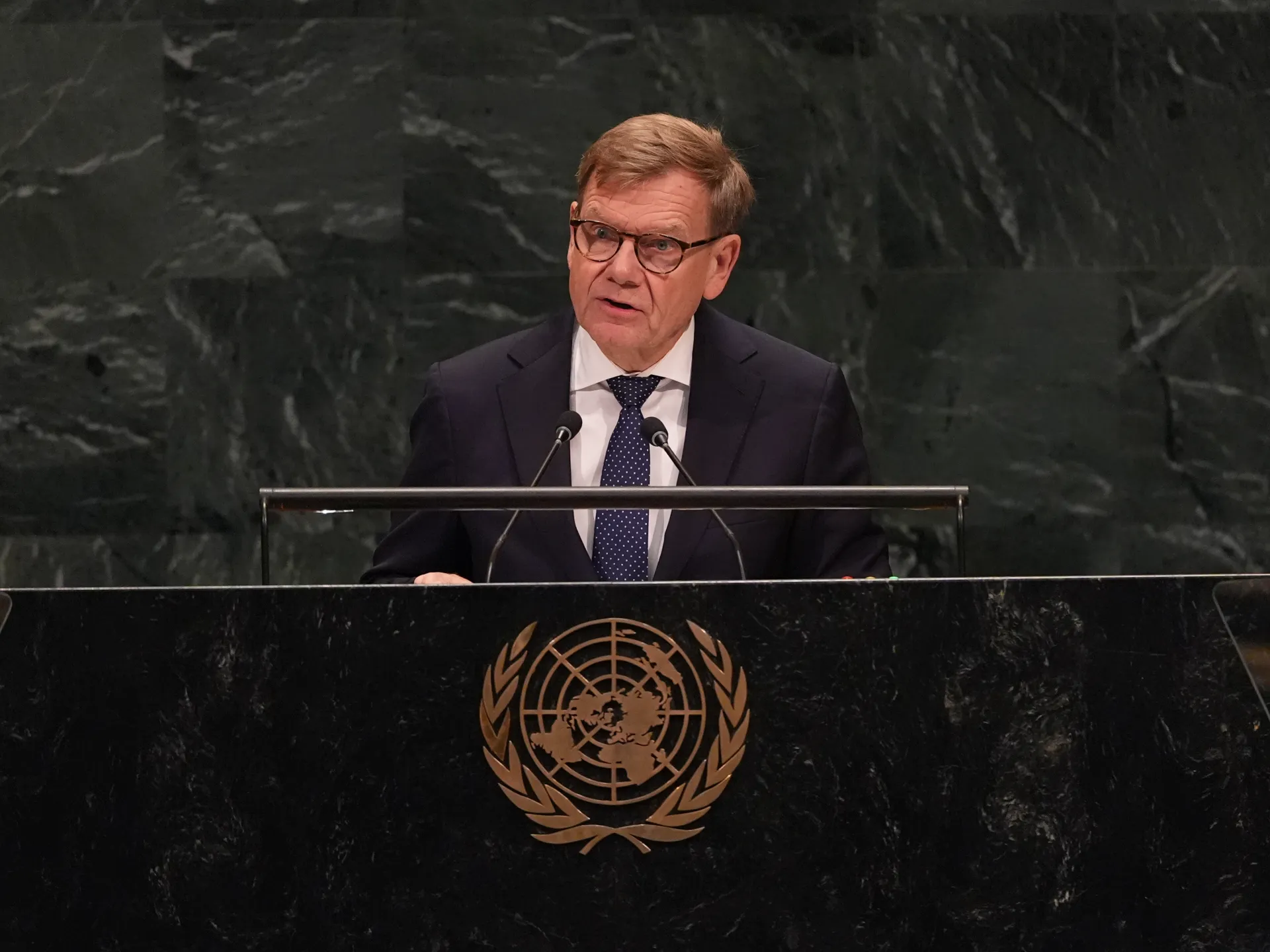 A suited man with glasses speaks at a lectern bearing the United Nations emblem.