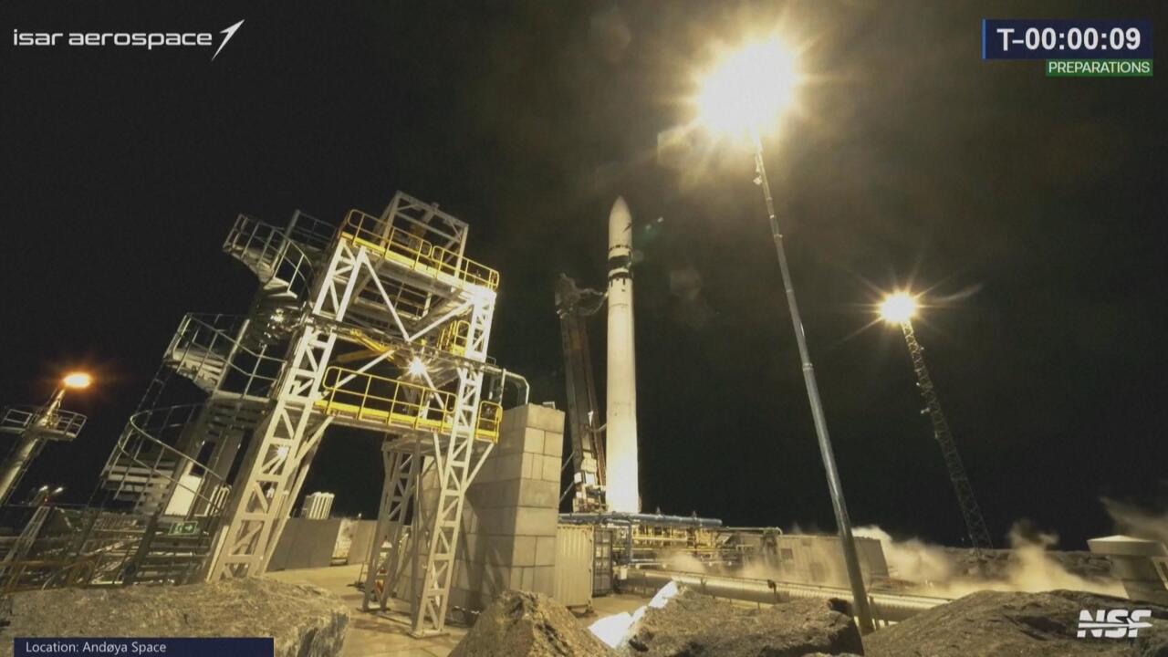 Rocket standing on a nighttime launch pad beside illuminated service structures and countdown graphics