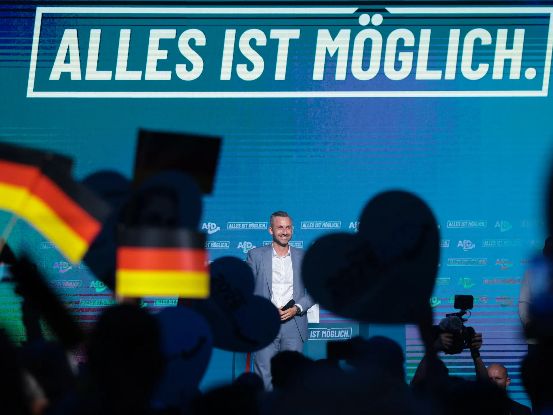 Man in suit stands onstage before a crowd holding German flags and cameras.