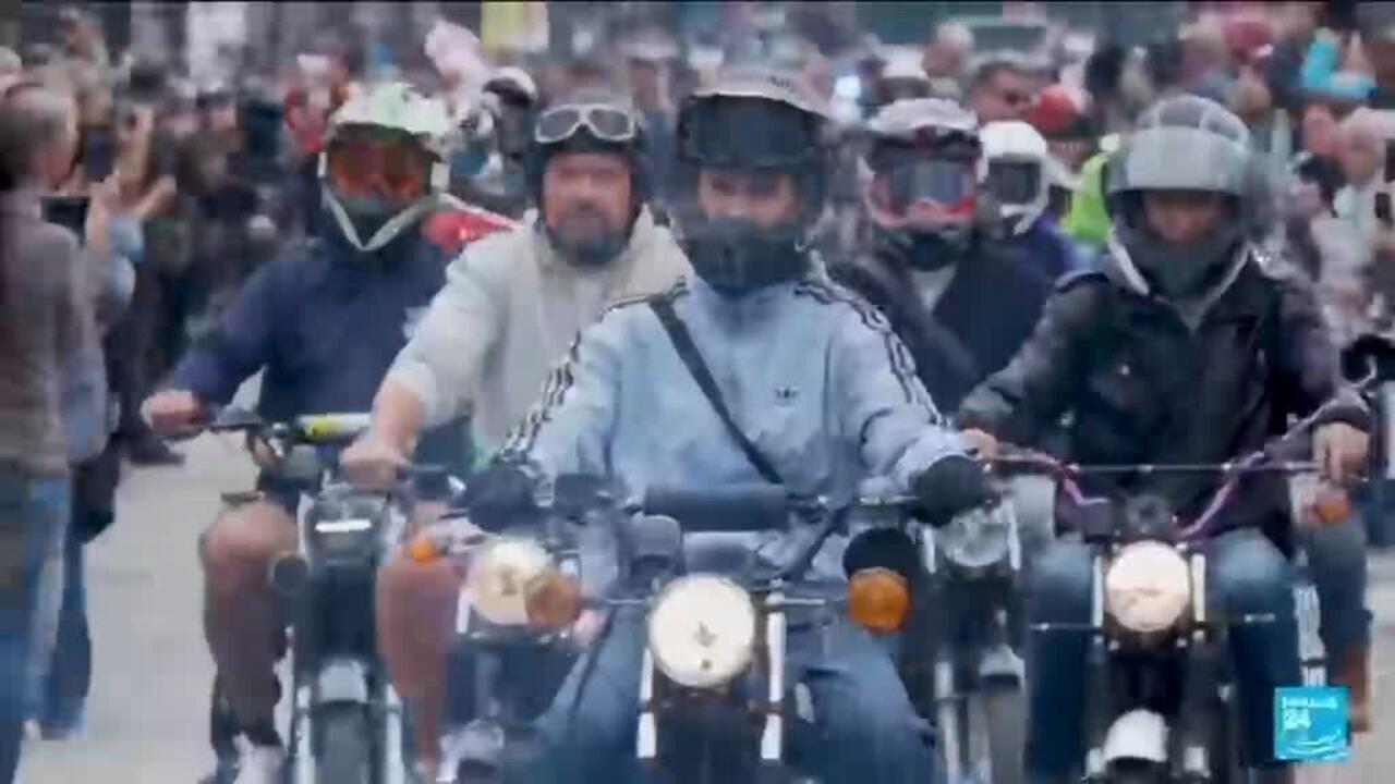 Helmeted motorcyclists ride closely together through a crowded outdoor gathering.