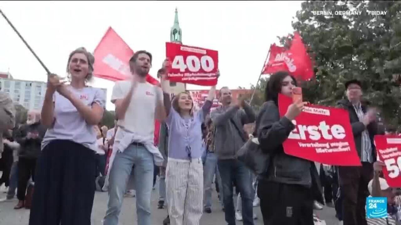 Demonstrators march outdoors holding red German-language signs and flags, with a church tower behind them.