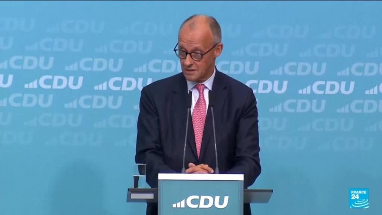 Bald man in glasses and pink tie speaking at a CDU-branded podium against blue backdrop