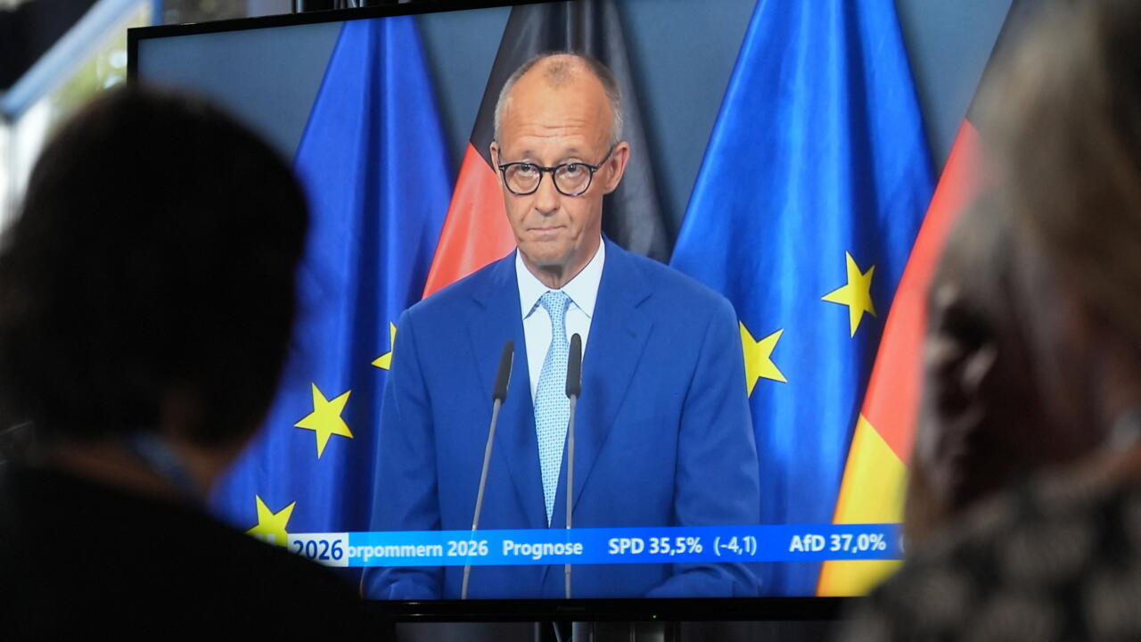 Man in blue suit speaking at a televised podium, framed by German and European Union flags