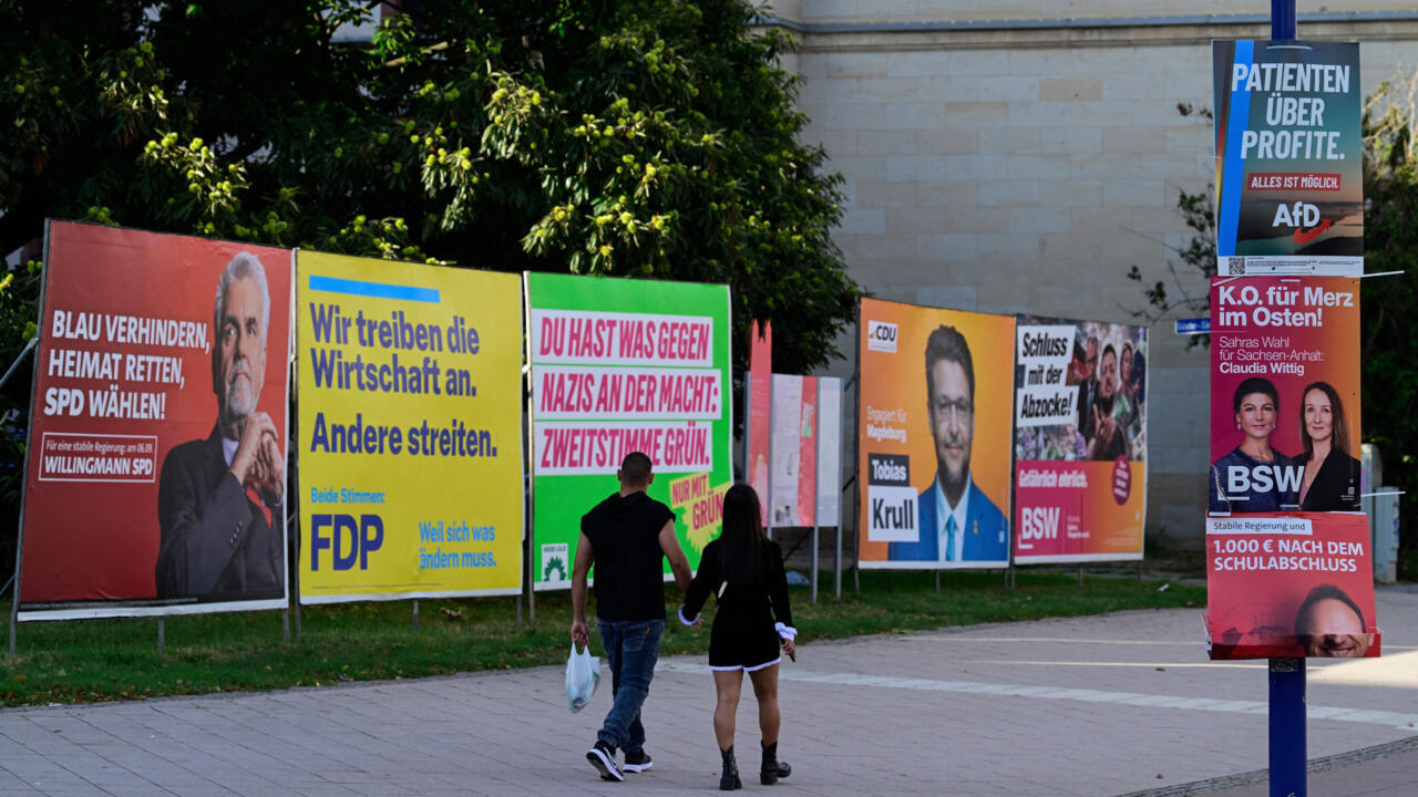 Pedestrians walk past colorful German political campaign posters displayed along a sidewalk.