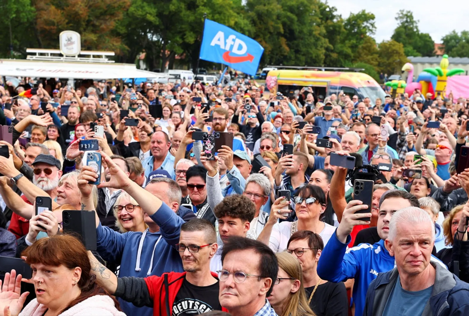 Large crowd holding smartphones beneath a blue AfD flag at an outdoor public gathering