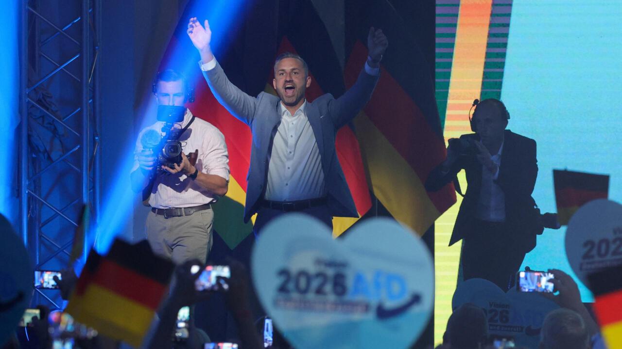 Man in suit raises arms on stage as supporters wave German flags and hold signs.