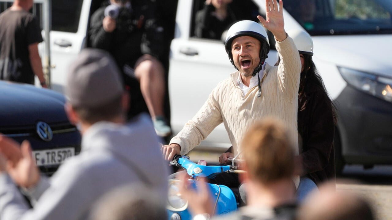 Helmeted man waves while riding a blue scooter through a crowd near parked vehicles.