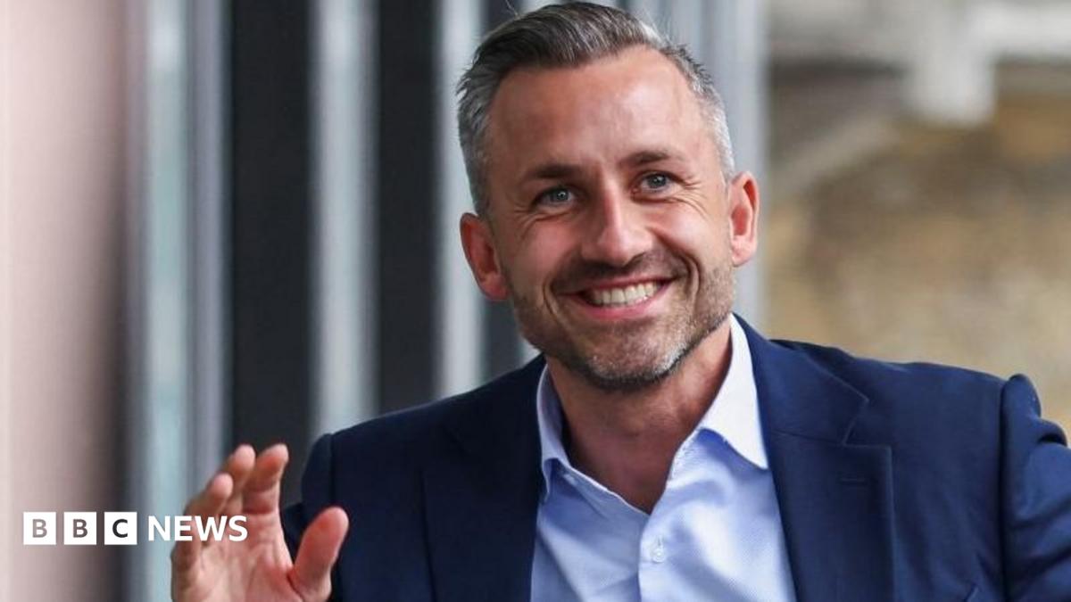 Smiling gray-haired man in a navy suit gesturing beside a window, with BBC News logo