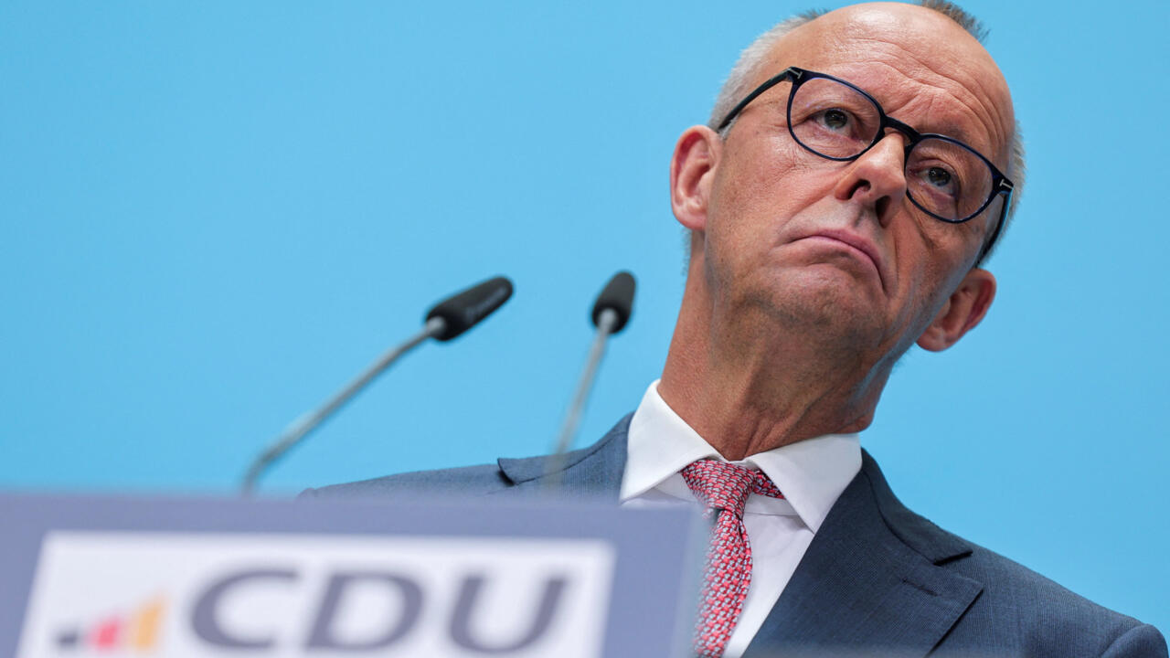 Man in glasses and suit speaking behind microphones against a blue backdrop