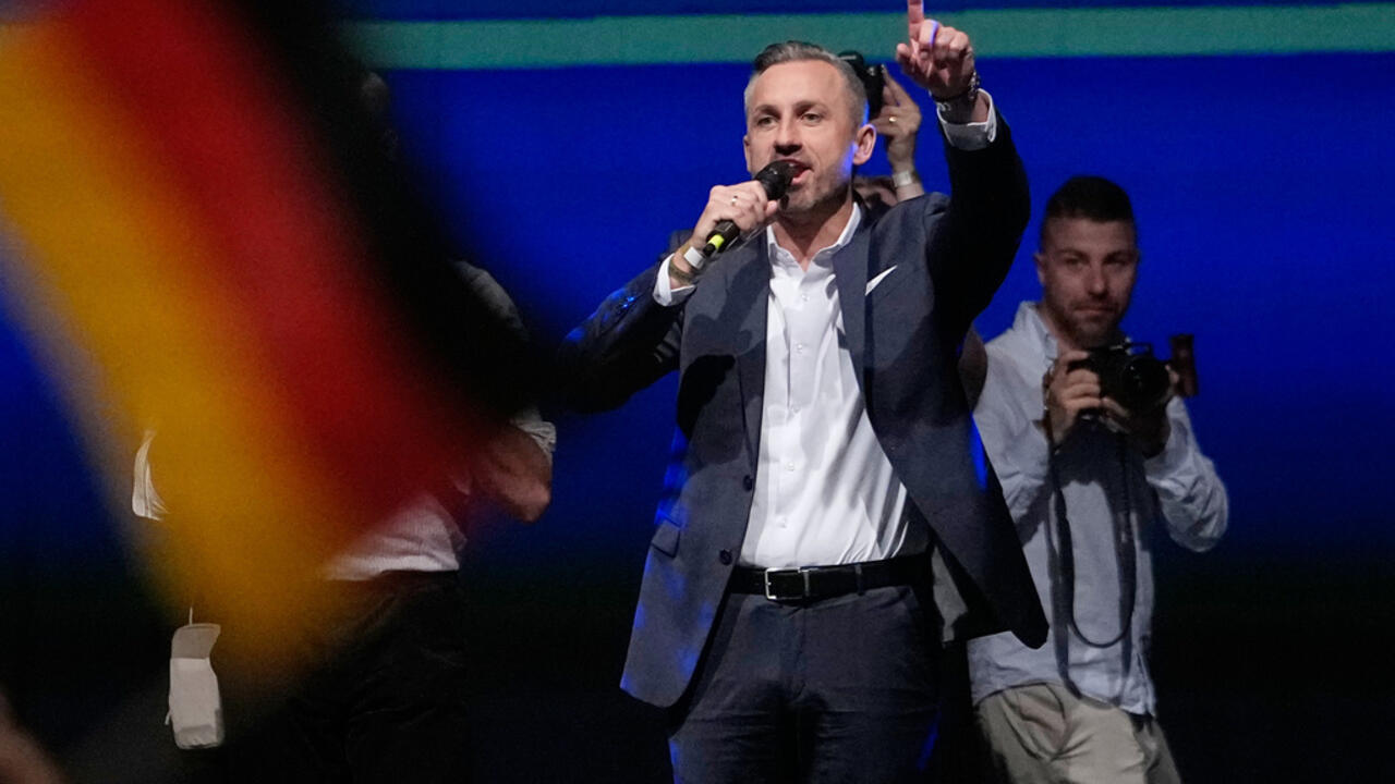 Man speaking into a microphone and raising one finger as a photographer stands behind him