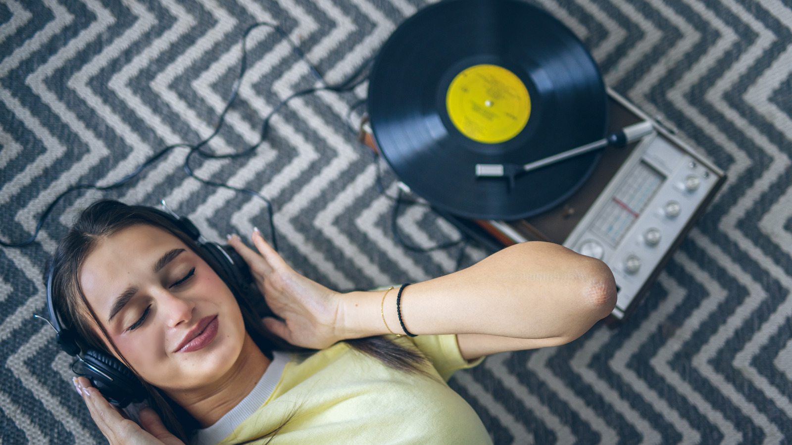 A woman listens through headphones beside a record player with a vinyl record on a patterned rug.