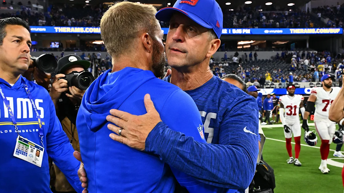 Two men in blue football apparel embrace on the field as photographers and players stand nearby.