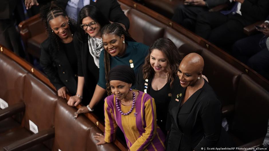 Five women stand together between wooden benches inside a legislative chamber.