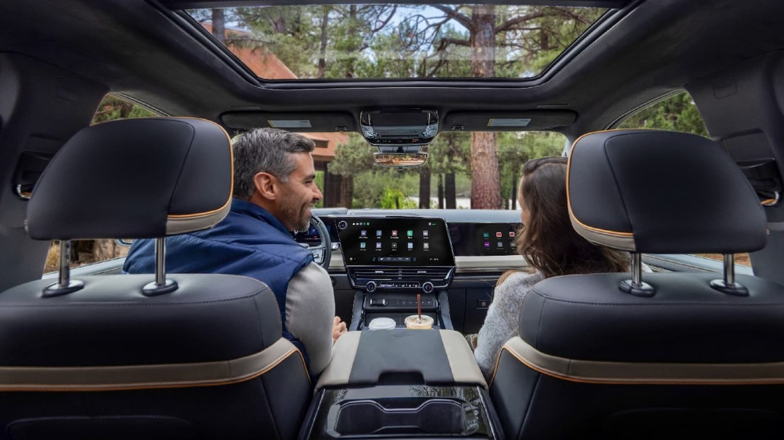View from the back seat of a car showing two occupants, dashboard screens, and panoramic sunroof.