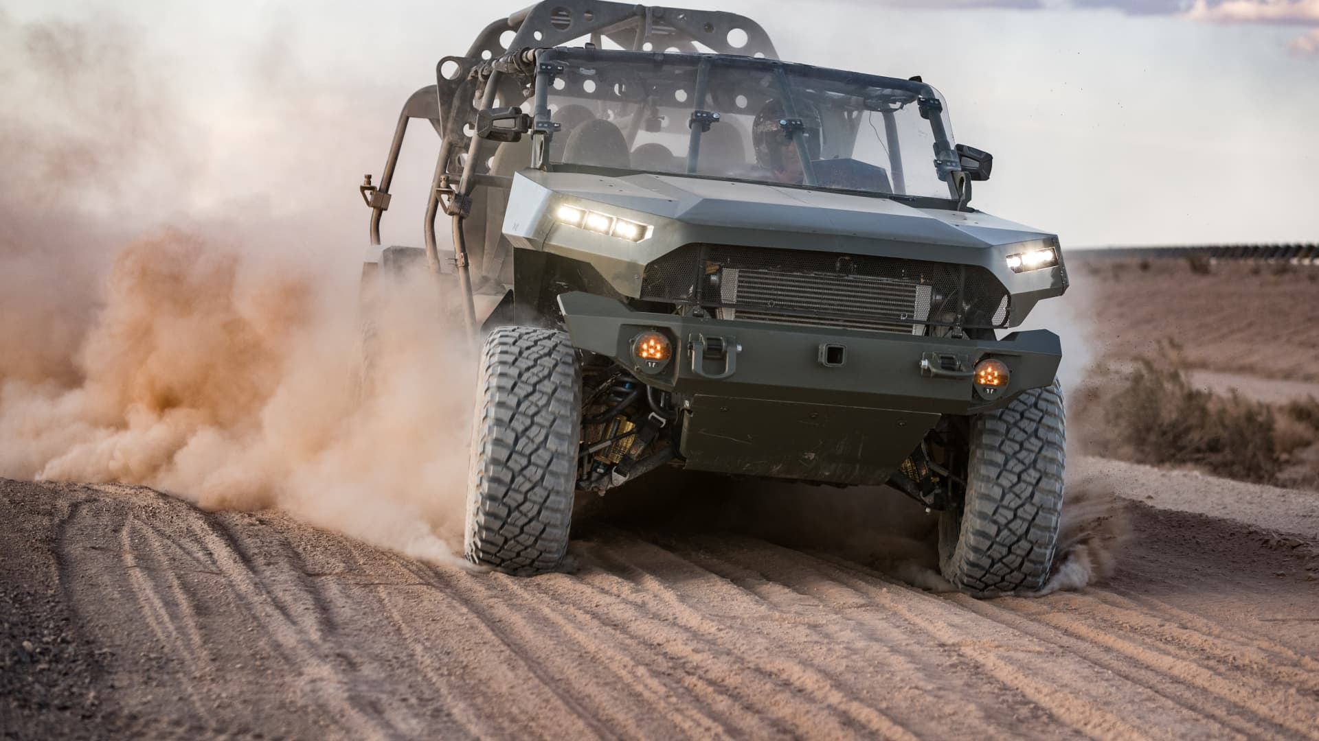 Off-road utility vehicle drives across a sandy track, raising a cloud of dust.